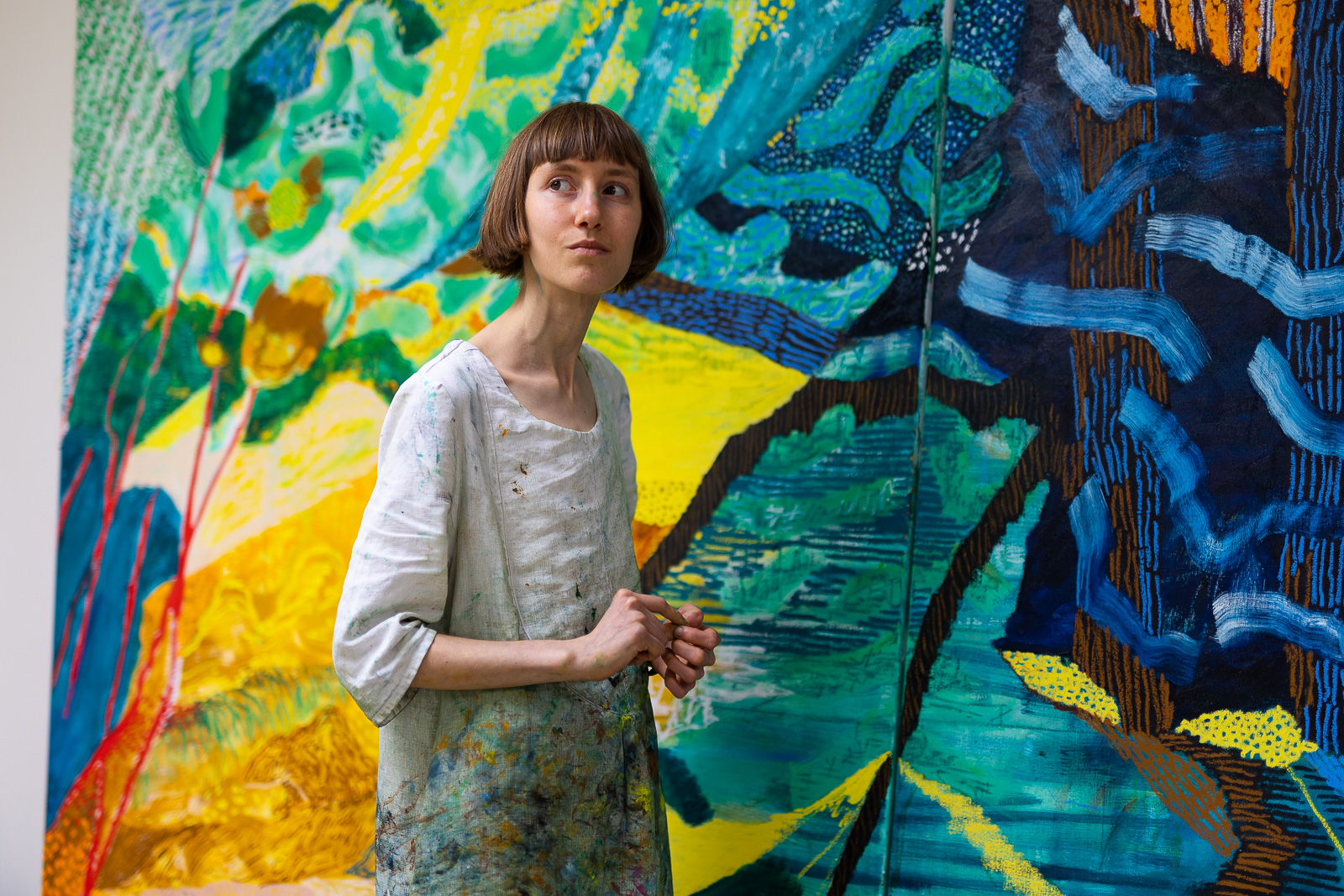 Anne Schillings staat in een tuniek met verfspetters voor een groot kleurrijk doek en kijkt bedachtzaam opzij